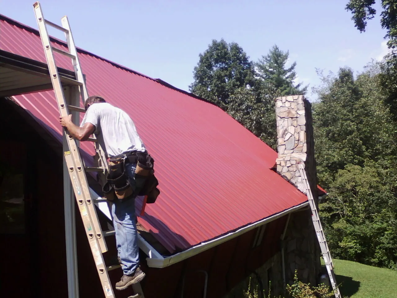 Roofer on a ladder beside a red metal roof and stone chimney — Soffit & Fascia Repair by Roofing Brothers in La Habra